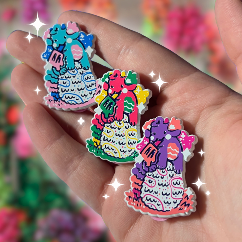 Three colorful dragon-shaped pins held in a hand with a blurred floral background