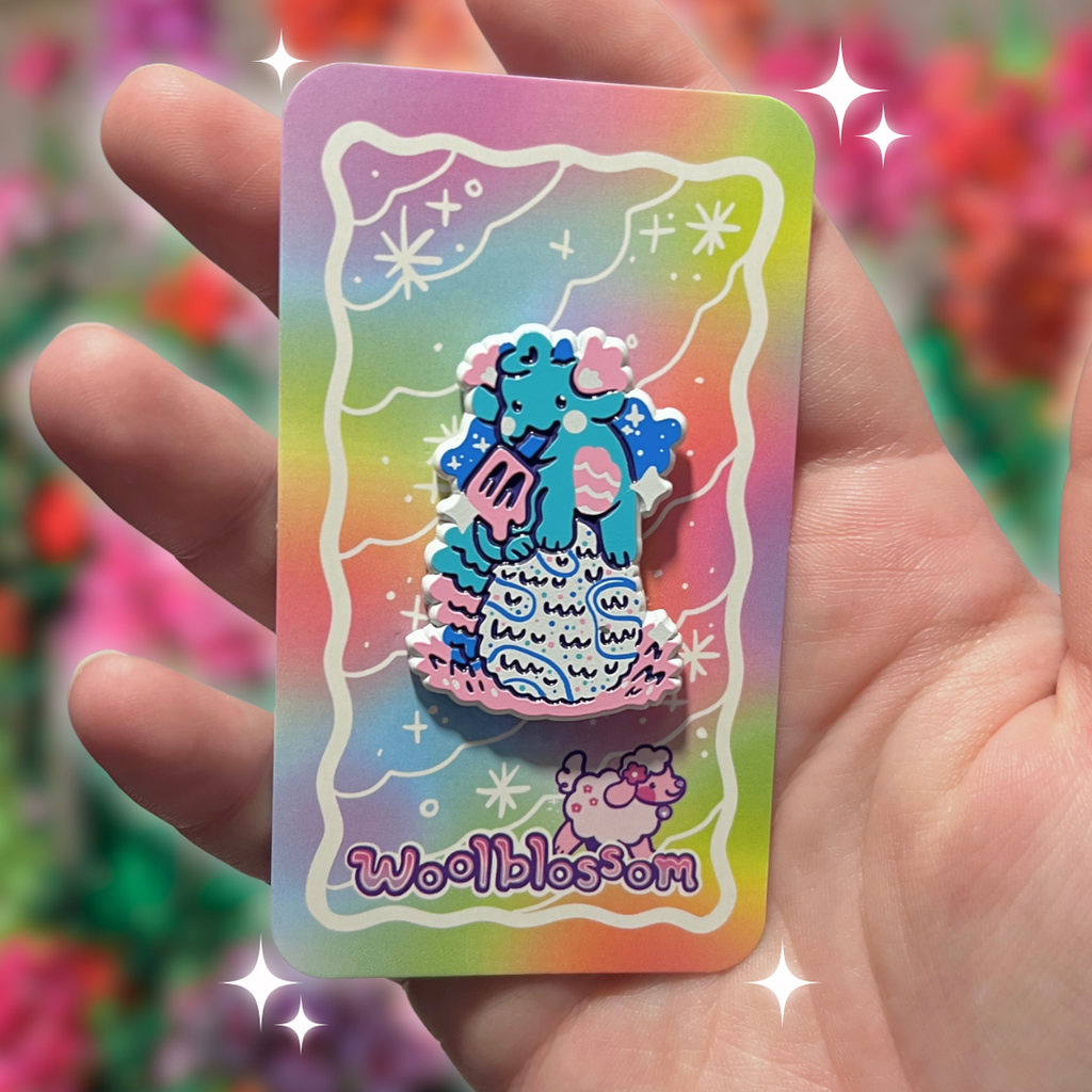 Hand holding a colorful card with a cartoon dragon enamel pin who is holding a popsicle and on top of an egg and 'Woolblossom' branding against a blurred floral background.