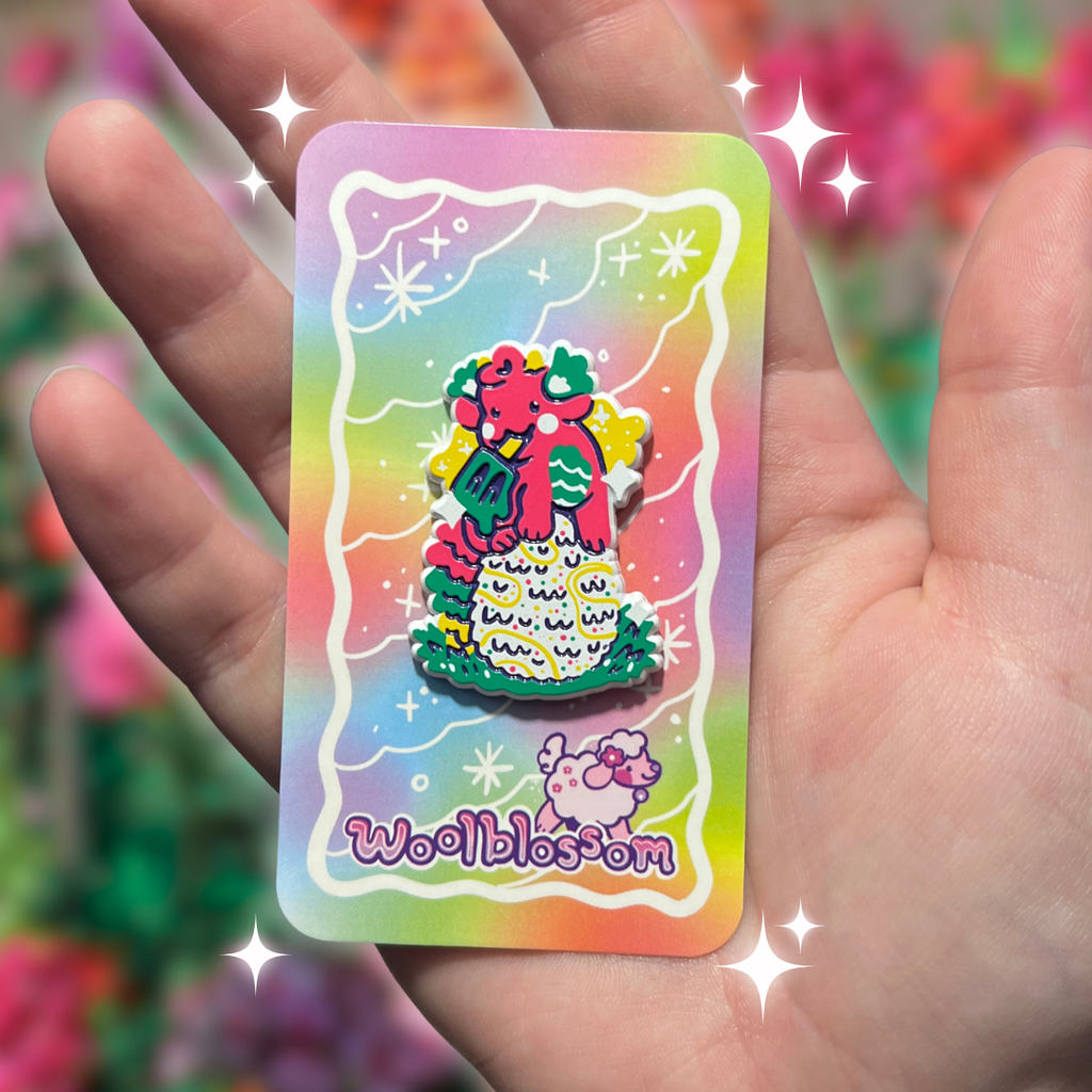 Hand holding a colorful card with a cartoon dragon enamel pin who is holding a popsicle and on top of an egg and 'Woolblossom' branding against a blurred floral background.
