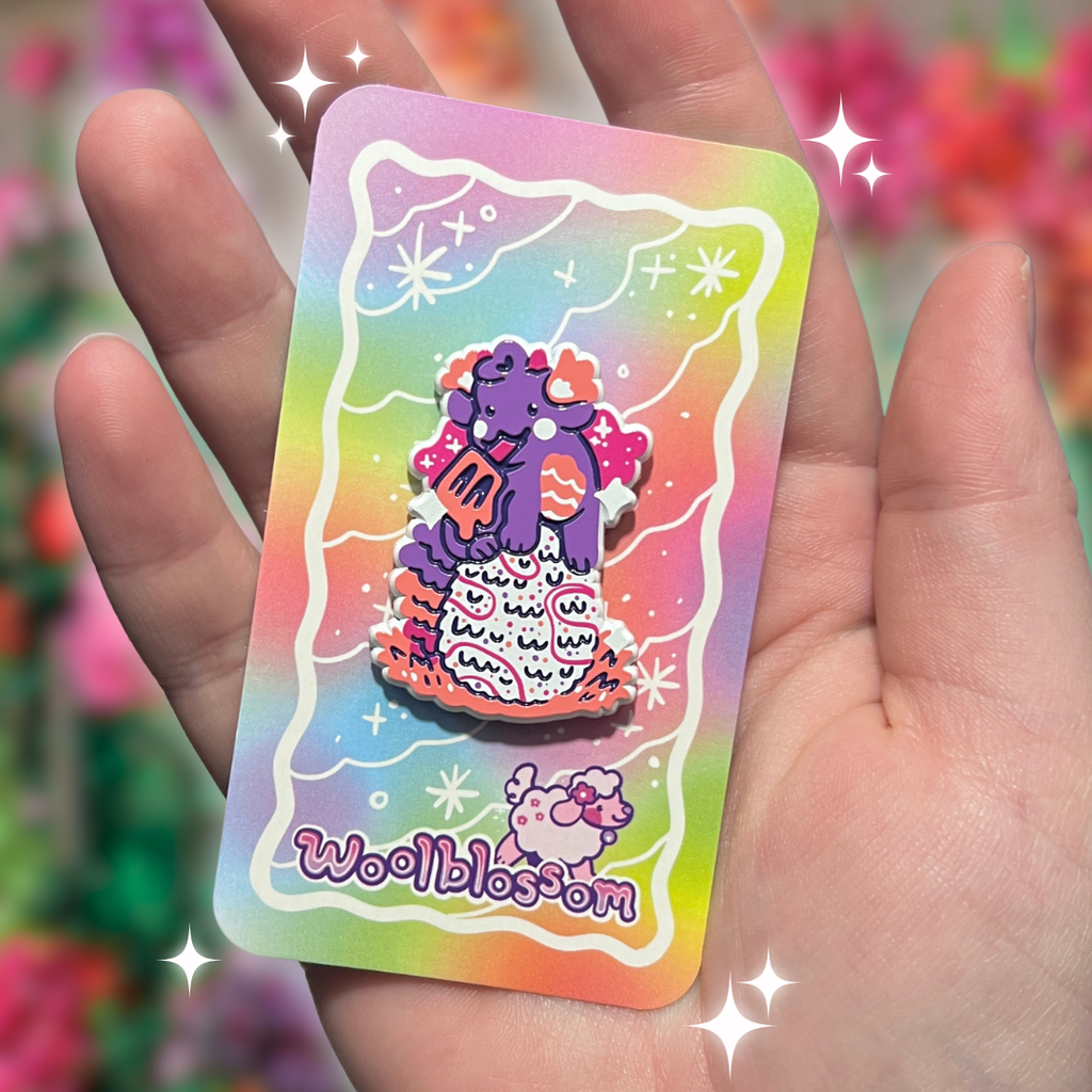 Hand holding a colorful card with a cartoon dragon enamel pin who is holding a popsicle and on top of an egg and 'Woolblossom' branding against a blurred floral background.