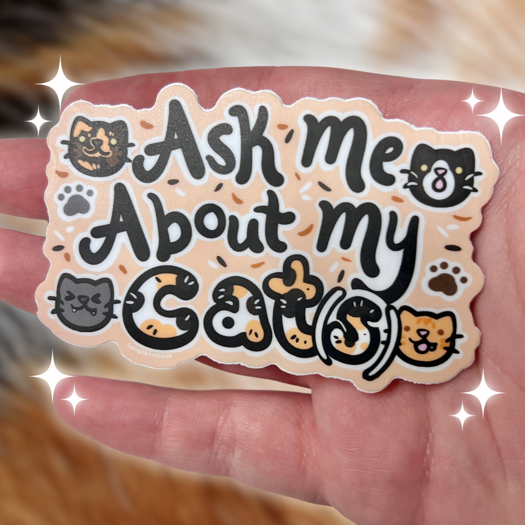 Sticker with 'Ask me About my Cats' text held in a hand, with a cat in the background.